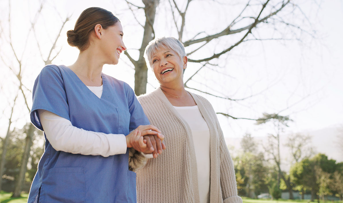 A smiling female healthcare professional walks arm in arm with an older smiling female patient